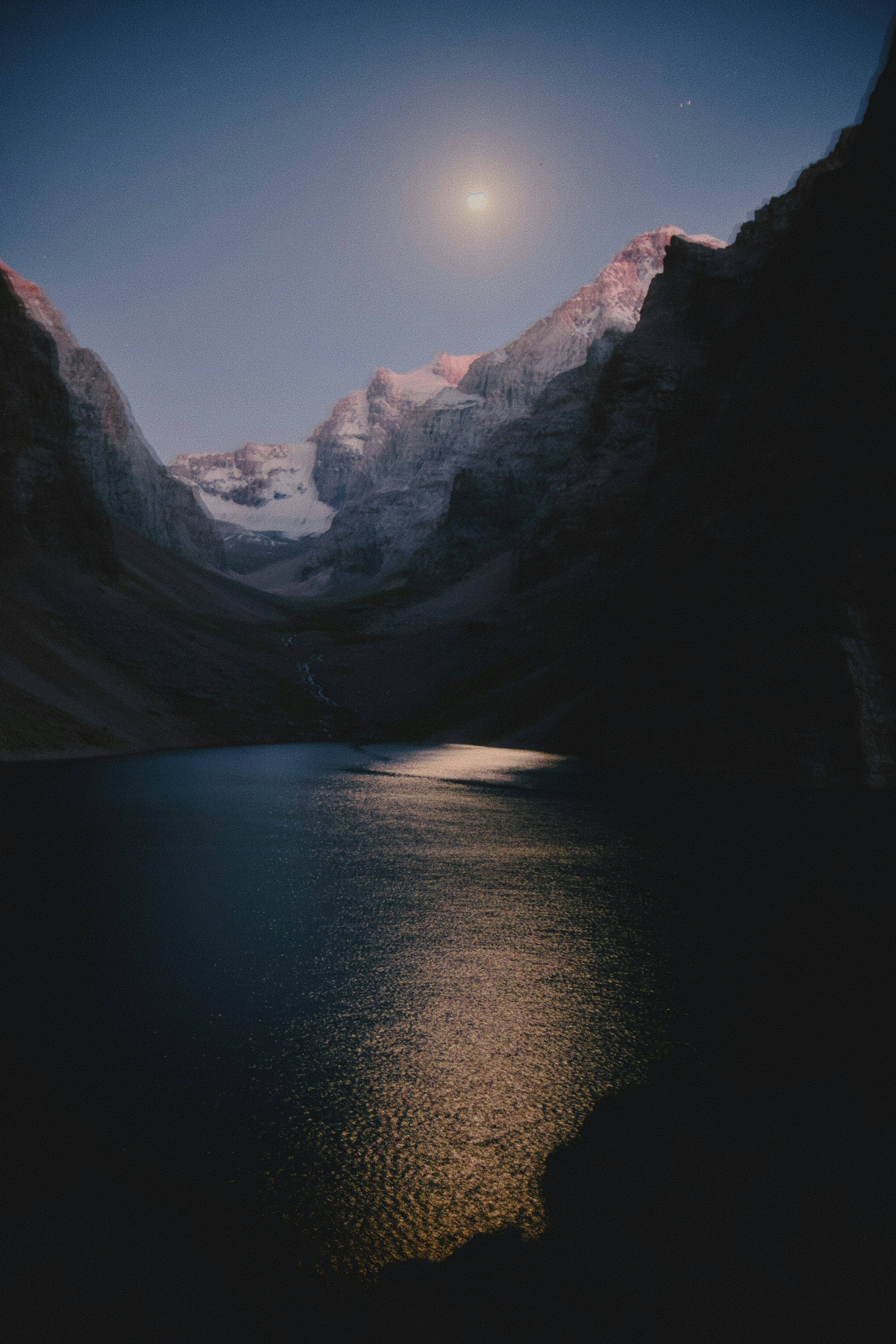 A full moon illuminating a dark mountain lake with rocky peaks in the distance